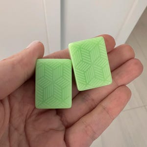 May include: Two light green rectangular tiles with a geometric cube pattern are held in a hand. The tiles have a glossy finish and a repeating design of interconnected shapes. The background is a plain white surface.
