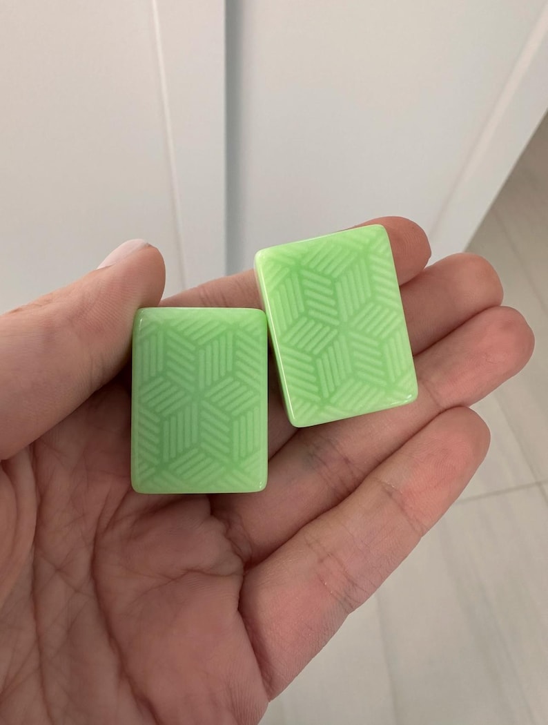 May include: Two light green rectangular tiles with a geometric cube pattern are held in a hand. The tiles have a glossy finish and a repeating design of interconnected shapes. The background is a plain white surface.