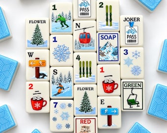 May include: A collection of rectangular mahjong tiles with winter-themed illustrations including skiers, snowflakes, a red cable car, a fireplace, and mugs of cocoa. The tiles feature text such as JOKER PASS, FLOWER, SOAP, RED, and GREEN.