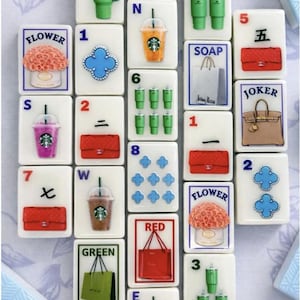 Puede incluir: Un conjunto de fichas de Mahjong blancas con varios dise&ntilde;os, incluyendo bolsos, flores, bebidas de Starbucks y n&uacute;meros. Las fichas est&aacute;n dispuestas sobre una superficie con estampado floral. Algunas fichas tienen las palabras "JOKER", "FLOWER", "SOAP", "RED" y "GREEN".
