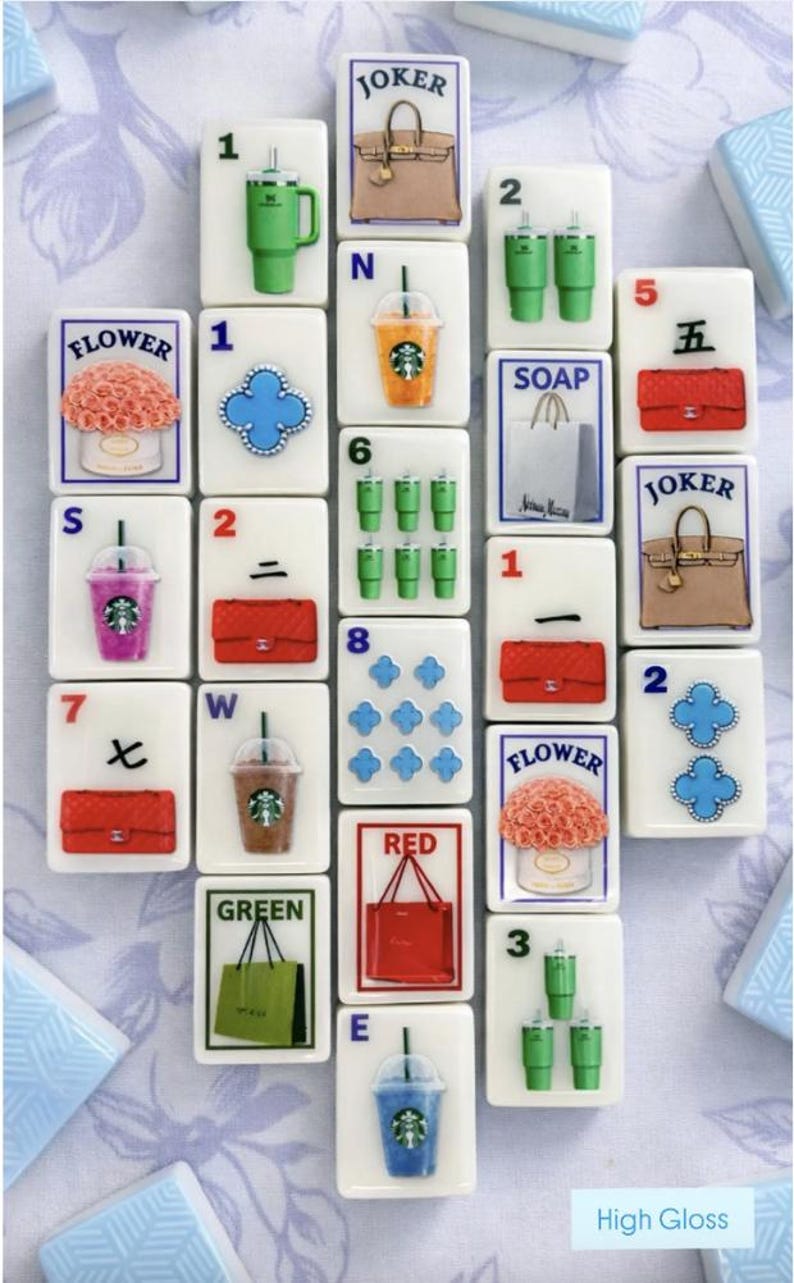 Puede incluir: Un conjunto de fichas de Mahjong blancas con varios dise&ntilde;os, incluyendo bolsos, flores, bebidas de Starbucks y n&uacute;meros. Las fichas est&aacute;n dispuestas sobre una superficie con estampado floral. Algunas fichas tienen las palabras "JOKER", "FLOWER", "SOAP", "RED" y "GREEN".