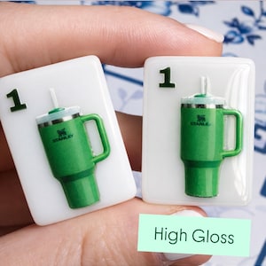Puede incluir: Dos baldosas rectangulares blancas con un dise&ntilde;o de vaso Stanley verde. Cada baldosa tiene el n&uacute;mero "1" en la esquina superior izquierda. El vaso es verde con un asa y una pajita blanca. El texto "High Gloss" est&aacute; en un rect&aacute;ngulo verde.