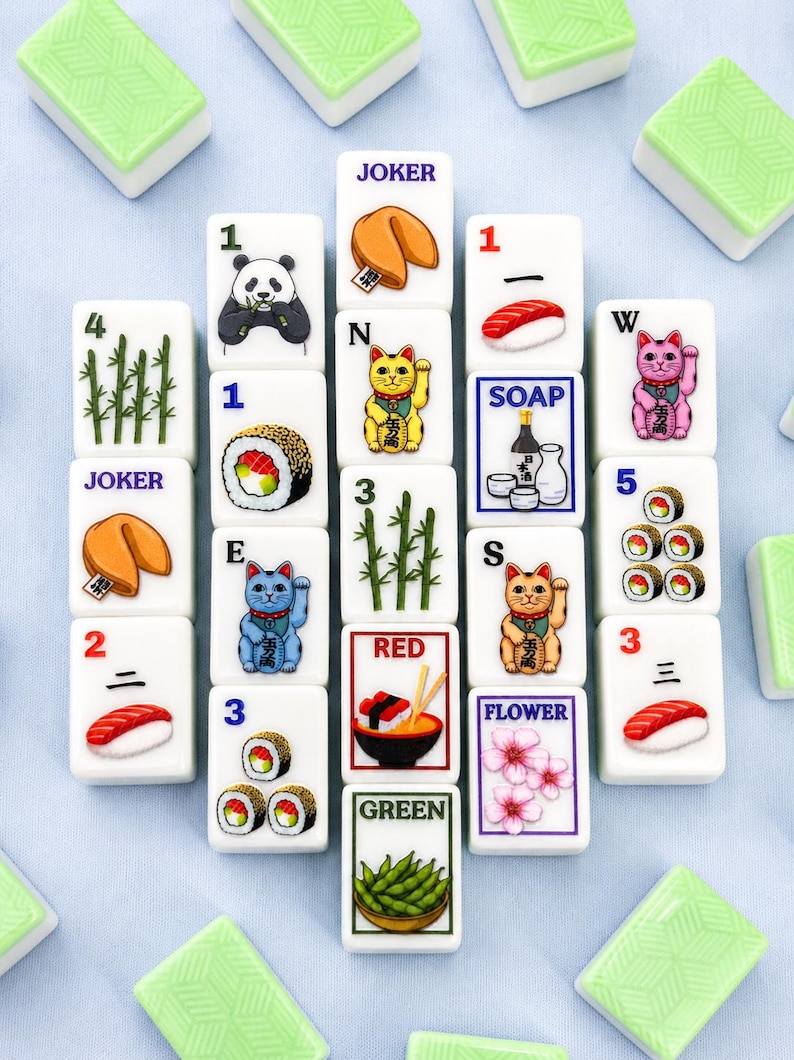 May include: A collection of white Mahjong tiles with various illustrations, including a panda, sushi, fortune cookies, and lucky cats. The tiles also feature numbers, Chinese characters, and the words "JOKER", "SOAP", "RED", "FLOWER", and "GREEN". Green tiles are scattered around the main set.