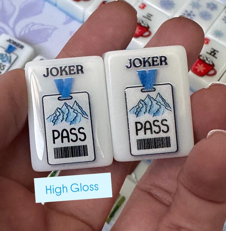May include: Two white, rectangular game tiles with a glossy finish. Each tile features the word "JOKER" above a blue pass with a mountain design and the word "PASS" below. A barcode is at the bottom. The tiles are held in a hand.