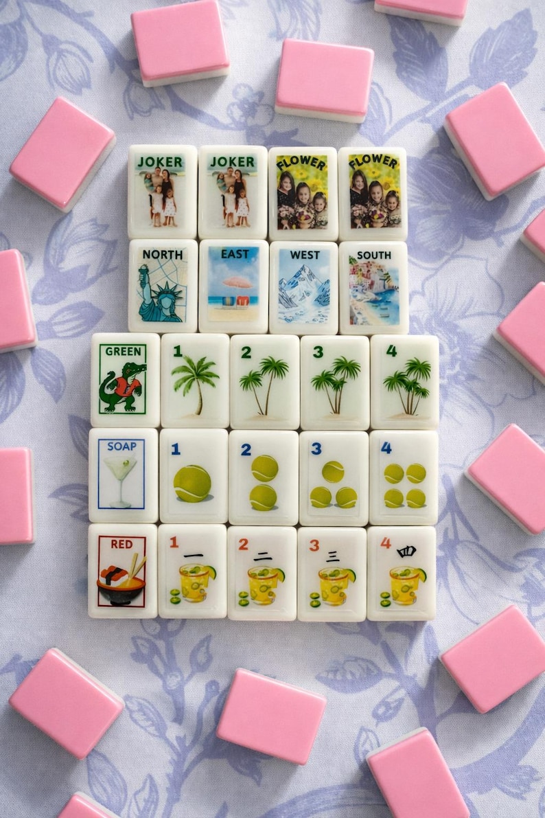 May include: A collection of white Mahjong tiles with various illustrations and text, including "JOKER," "FLOWER," and directional markers. Pink tiles surround the set, arranged on a floral patterned surface.