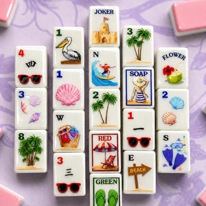 May include: A collection of white mahjong tiles with beach-themed illustrations, including a pelican, palm trees, sunglasses, and a sandcastle. The tiles are arranged on a purple surface, with pink tiles in the background.