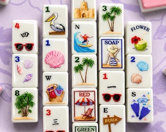 May include: A collection of white mahjong tiles with beach-themed illustrations, including a pelican, palm trees, sunglasses, and a sandcastle. The tiles are arranged on a purple surface, with pink tiles in the background.