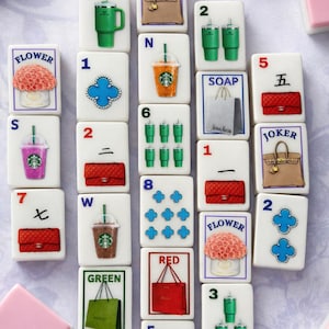 Puede incluir: Un conjunto de fichas de Mahjong blancas con varias ilustraciones, incluyendo bolsos, flores y bebidas. Las fichas presentan n&uacute;meros, letras y s&iacute;mbolos, con fichas rectangulares rosas y blancas.