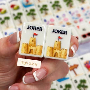 May include: Two white Mahjong tiles with a sandcastle design and the word "JOKER" in black. The sandcastle is yellow with a red flag. The tiles are held in front of other tiles with various beach-themed images.