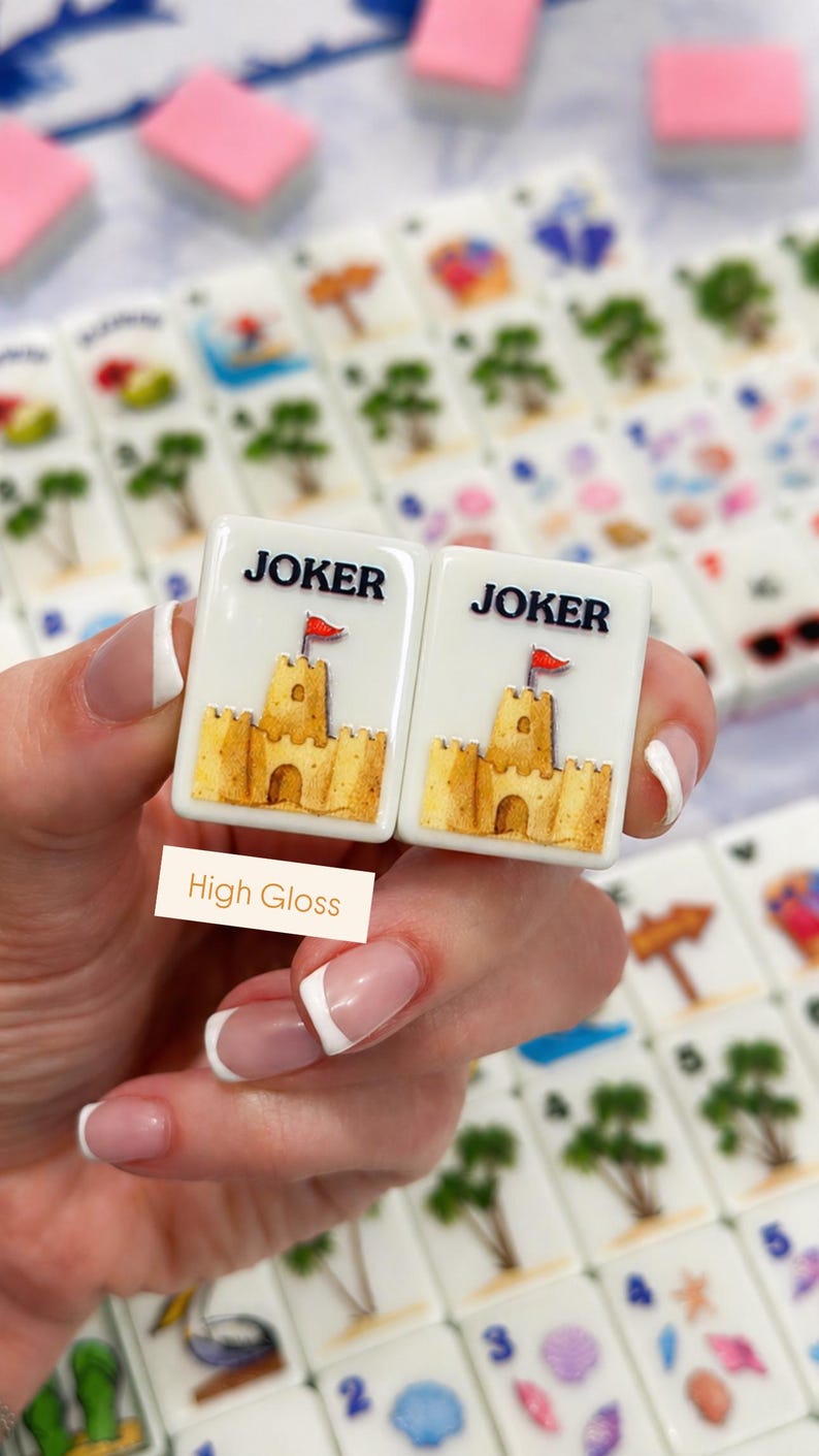 May include: Two white Mahjong tiles with a sandcastle design and the word "JOKER" in black. The sandcastle is yellow with a red flag. The tiles are held in front of other tiles with various beach-themed images.