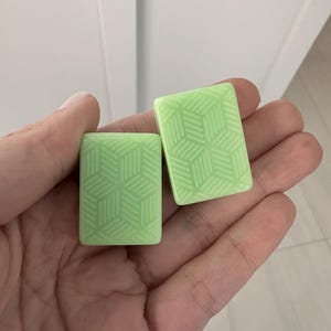 K&ouml;nnte beinhalten: Zwei hellgr&uuml;ne rechteckige Kacheln mit einem geometrischen W&uuml;rfelmuster werden in einer Hand gehalten. Die Kacheln haben eine gl&auml;nzende Oberfl&auml;che und ein sich wiederholendes Design aus ineinandergreifenden W&uuml;rfeln. Der Hintergrund ist eine schlichte wei&szlig;e Wand.