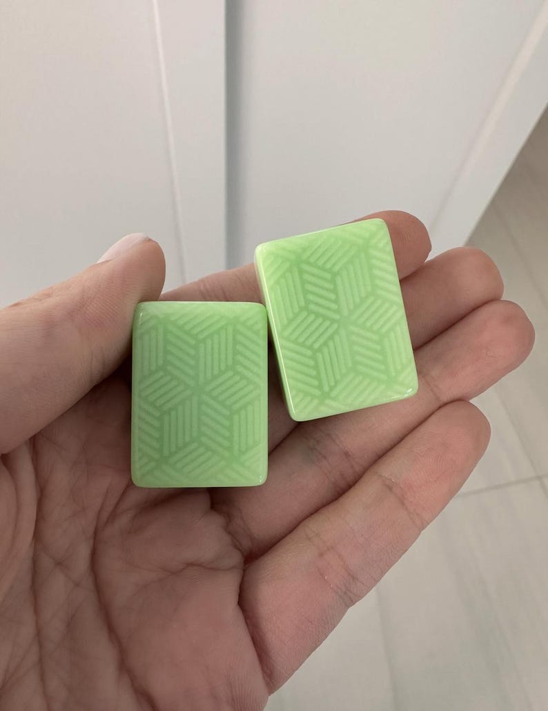 K&ouml;nnte beinhalten: Zwei hellgr&uuml;ne rechteckige Kacheln mit einem geometrischen W&uuml;rfelmuster werden in einer Hand gehalten. Die Kacheln haben eine gl&auml;nzende Oberfl&auml;che und ein sich wiederholendes Design aus ineinandergreifenden W&uuml;rfeln. Der Hintergrund ist eine schlichte wei&szlig;e Wand.