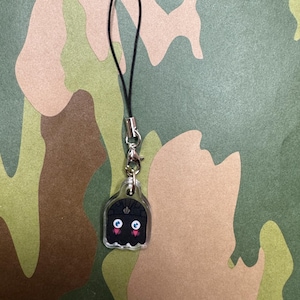 May include: A black and white ghost charm with pink cheeks and blue eyes. The charm is attached to a black string.