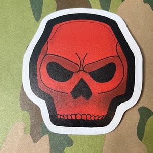 May include: A red skull sticker with a black outline. The skull has a menacing expression with furrowed brows and a frown.