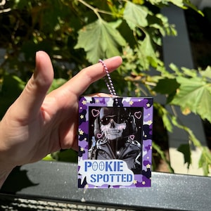 May include: A purple and black keychain with a skull wearing sunglasses and a heart-shaped frame. The keychain has the text "POOKIE SPOTTED" on it.