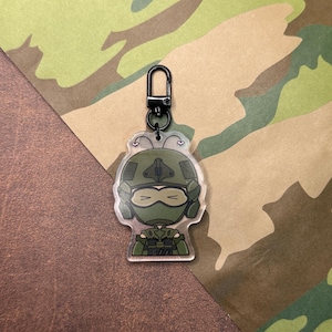 May include: A green and black cartoon character keychain with a black metal clasp. The character is wearing a green helmet and a green and black camouflage uniform.
