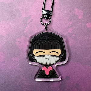 May include: A black and white cartoon character keychain with a pink heart. The character is wearing a black beanie and a white mask. The keychain has a black metal clasp.