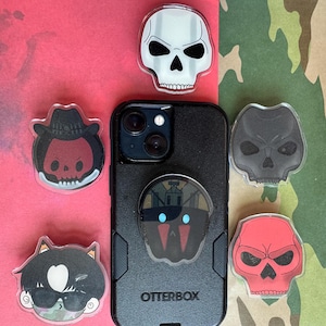 May include: A black Otterbox phone case with a pop-up grip featuring a skull design. Several acrylic skull-themed phone grips surround the phone, in various colours and styles, including a cat, a cowboy, and a red skull.