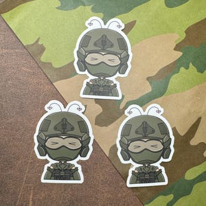 May include: Three cartoon stickers of a person wearing a green helmet and camouflage clothing. The stickers have pink hearts on top of the helmet.