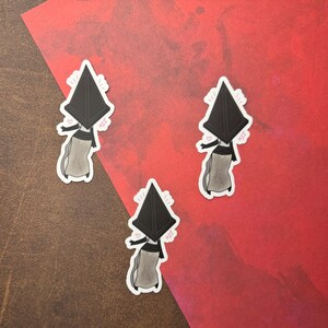 May include: Three black and white stickers of a cartoon character wearing a pyramid-shaped head. The character is wearing a long, flowing dress and has a small, smiling face. The stickers are on a red background.