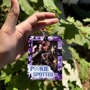 May include: A purple and white camouflage-patterned photo frame with a picture of a person wearing a camouflage hat and holding a gun. The frame has a white border and the text "POOKIE SPOTTED" in blue letters.