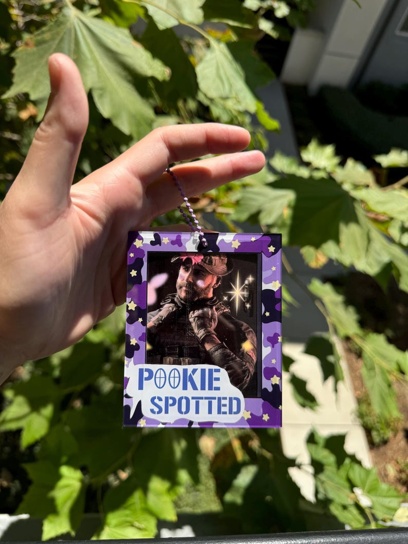 May include: A purple and white camouflage-patterned photo frame with a picture of a person wearing a camouflage hat and holding a gun. The frame has a white border and the text "POOKIE SPOTTED" in blue letters.