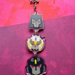 May include: A keychain featuring three cartoon robot heads. The top head is grey with red eyes, the middle head is white with yellow and red accents and red eyes, and the bottom head is dark blue with yellow accents and red eyes.