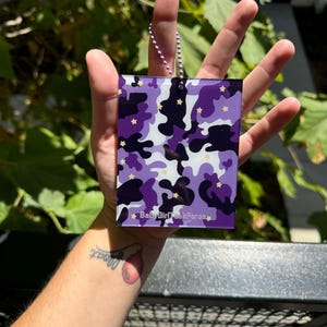 May include: Purple and white camouflage patterned keychain with gold stars and the text "Baby Girl Task Force" on a silver chain.