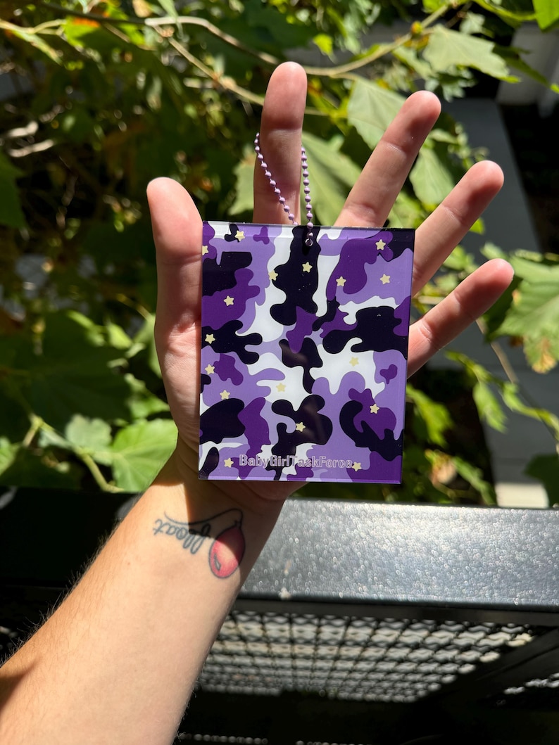 May include: Purple and white camouflage patterned keychain with gold stars and the text "Baby Girl Task Force" on a silver chain.