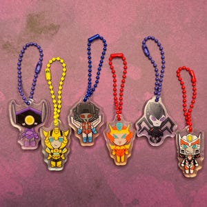 May include: Seven colourful keychains featuring cartoon characters from the Transformers franchise. The characters are Optimus Prime, Megatron, Bumblebee, Starscream, Soundwave, and two others. The keychains are made of clear plastic with a silver chain.