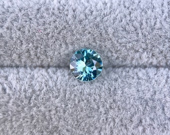 Può includere: Una pietra preziosa rotonda di colore blu chiaro con taglio a faccette, posizionata su uno sfondo grigio e soffice. Il testo "Under Shade" è visibile nell'angolo in alto a sinistra dell'immagine.