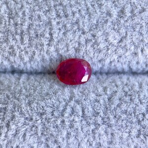 May include: A vibrant red oval-shaped gemstone, likely a ruby, is displayed on a grey, fuzzy surface. The text "Under Shade" is visible in the top left corner of the image.