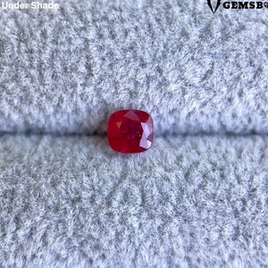 May include: A deep red cushion-cut gemstone on a gray fabric background. The gemstone is a ruby.