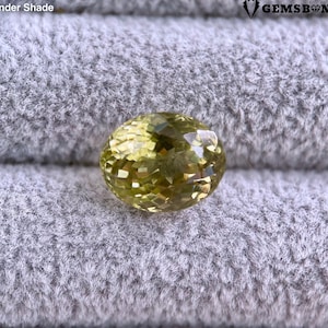 Puede incluir: Una gema facetada de color amarillo verdoso y forma ovalada. La gema se encuentra sobre una tela gris texturizada. La imagen incluye el texto "Under Shade" y el logotipo de Gemsbond.