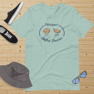 May include: A light blue t-shirt with a graphic of two Michigan pastries, a paczki and a pastie, with the text "Michigan's Stuffed Pastries".