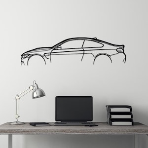 May include: A black metal wireframe silhouette of a sports car mounted on a white wall above a desk with a laptop, lamp, and a black stool.