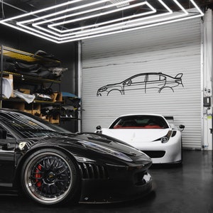 May include: Two sports cars, one black and one white, are parked in a garage. The black car has a red brake caliper. The garage features a white roller door with a car outline and a geometric light fixture overhead. The floor is black.