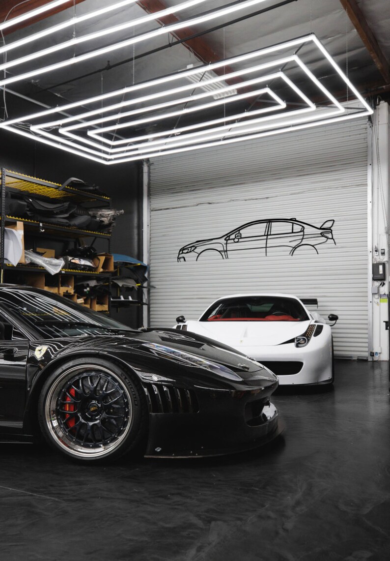 May include: Two sports cars, one black and one white, are parked in a garage. The black car has a red brake caliper. The garage features a white roller door with a car outline and a geometric light fixture overhead. The floor is black.