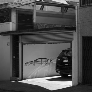 May include: Monochrome image of a garage with a car inside. A car outline is drawn on the wall. The garage door is partially open, revealing the rear of a car. The building has a flat roof.