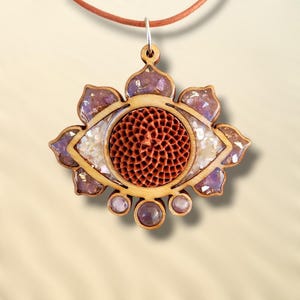 Puede incluir: Collar con colgante de madera con diseño de ojo. El colgante tiene un marco de madera marrón claro con detalles morados y blancos. El centro del ojo es un círculo texturizado de color marrón rojizo. El collar tiene un cordón de cuero marrón.