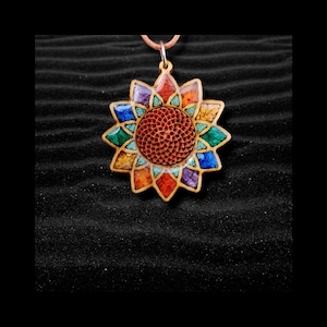 May include: A wooden pendant necklace with a sunflower design. The pendant features a central, textured, reddish-brown circle surrounded by colorful, geometric shapes in shades of orange, blue, green, purple, and yellow. The necklace has a brown leather cord.