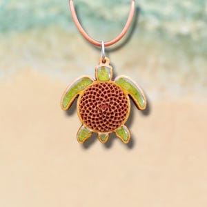 Puede incluir: Colgante de tortuga de madera con un centro marrón estampado y detalles verdes. La tortuga está suspendida de un cordón de cuero marrón. El fondo presenta una imagen borrosa de una playa y el océano.