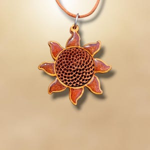 Puede incluir: Un colgante de madera con forma de girasol con un cordón marrón. El colgante tiene un centro detallado y texturizado y bordes en forma de pétalos en tonos marrones y naranjas. El colgante está suspendido de un cordón fino de color marrón claro.