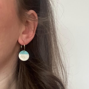 Puede incluir: Primer plano de un pendiente de aro dorado con un colgante redondo de cerámica. El colgante está dividido en dos partes: una base blanca moteada y una sección superior azul claro. El pendiente se muestra sobre un fondo de cabello castaño y una pared blanca.