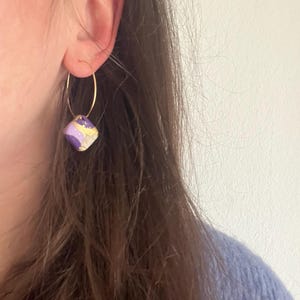 Könnte beinhalten: Goldener Creolen-Ohrring mit einem quadratischen, lila-goldenen Anhänger. Der Anhänger hat ein marmoriertes Aussehen mit goldenen Akzenten. Der Ohrring wird am Ohr getragen, der Hintergrund ist unscharf.