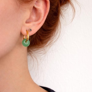 May include: Gold hoop earrings with a round, green stone pendant. The earrings are worn on a person's ear, with a small stud earring visible above the hoop. The background is a plain white wall.