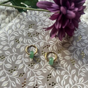 May include: A pair of gold hoop earrings with a small, light green, cylindrical bead hanging from each. The earrings are displayed on a decorative, light gray surface with a floral pattern, and a purple flower and green leaf are in the background.