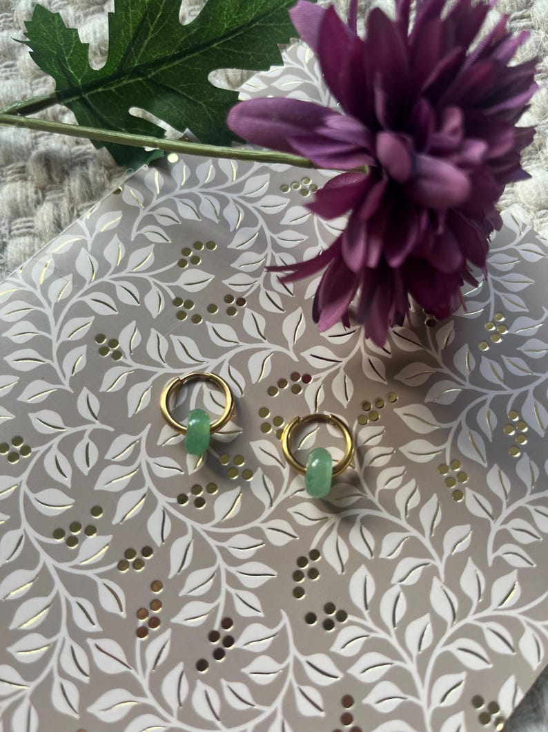 May include: A pair of gold hoop earrings with a small, light green, cylindrical bead hanging from each. The earrings are displayed on a decorative, light gray surface with a floral pattern, and a purple flower and green leaf are in the background.