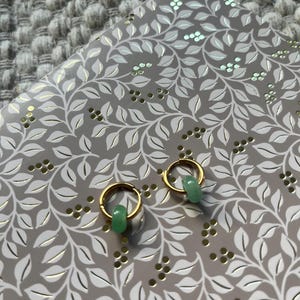 May include: A pair of gold hoop earrings with small, light green jade beads. The earrings are displayed on a gray notebook with a white and gold leaf pattern. The background is a textured, gray surface.