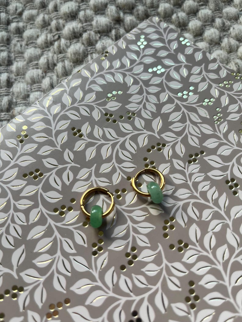 May include: A pair of gold hoop earrings with small, light green jade beads. The earrings are displayed on a gray notebook with a white and gold leaf pattern. The background is a textured, gray surface.